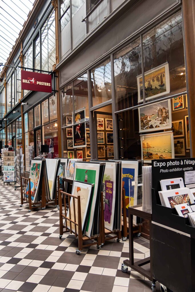 Photo d’une galerie commerciale couverte : boutique d’affiches avec cadres et posters exposés en vitrine et sur des présentoirs roulants, sol en damier noir et blanc, verrière au-dessus.