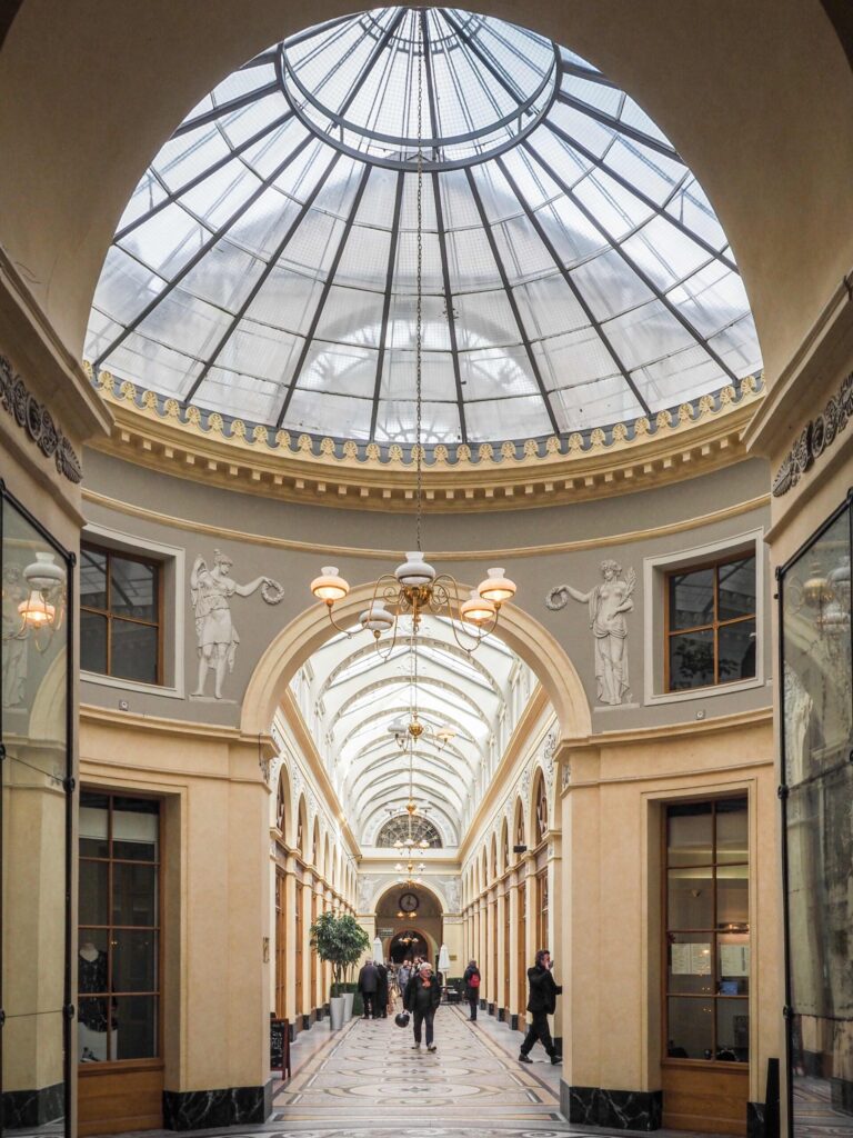 Photo en perspective d’un passage couvert, Galerie Vivienne Paris : verrière en coupole, décor classique avec statues murales, arcades et sols mosaïqués, lampes suspendues et personnes marchant au centre.