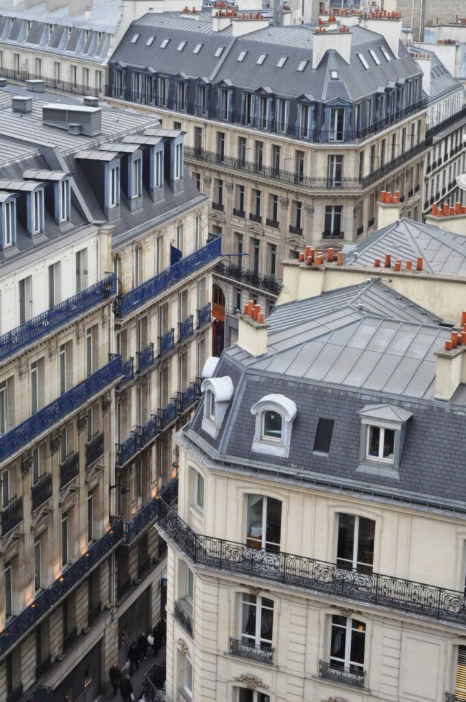 enchevêtrement d’immeubles haussmanniens aux façades claires, toits en zinc gris, rangées de fenêtres, balcons en fer forgé et nombreuses cheminées, dans une rue étroite en contrebas.