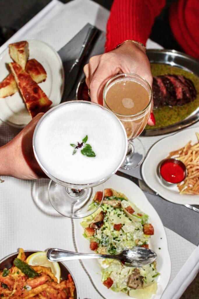 Vue du dessus d’une table de restaurant : deux verres portés pour trinquer (cocktail blanc mousseux avec feuille verte, boisson ambrée), entourés d’une salade, de frites avec sauce rouge, de pain et d’un plat de viande.