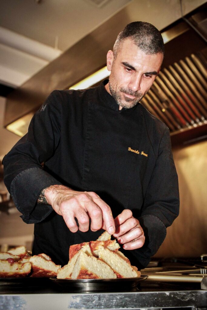 Photo en cuisine : un chef en veste noire, concentré, émiette/ajuste des tranches de pain sur un plat, avec la hotte et les équipements inox en arrière-plan.