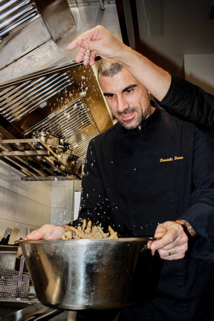 Photo en cuisine : un chef en veste noire (prénom brodé) sale un plat, les grains tombent en pluie au-dessus d’une grande casserole remplie de pâtes, avec hotte et équipements inox en arrière-plan.