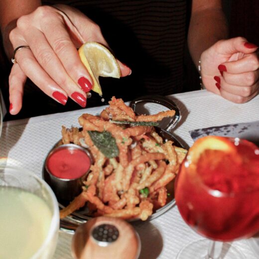 Photo à table : deux personnes, une main aux ongles rouges presse un quartier de citron sur une assiette de fritures croustillantes garnies d’herbes, avec ramequin de sauce rouge, sa-lière/poivrière et un verre de cocktail rouge au premier plan.