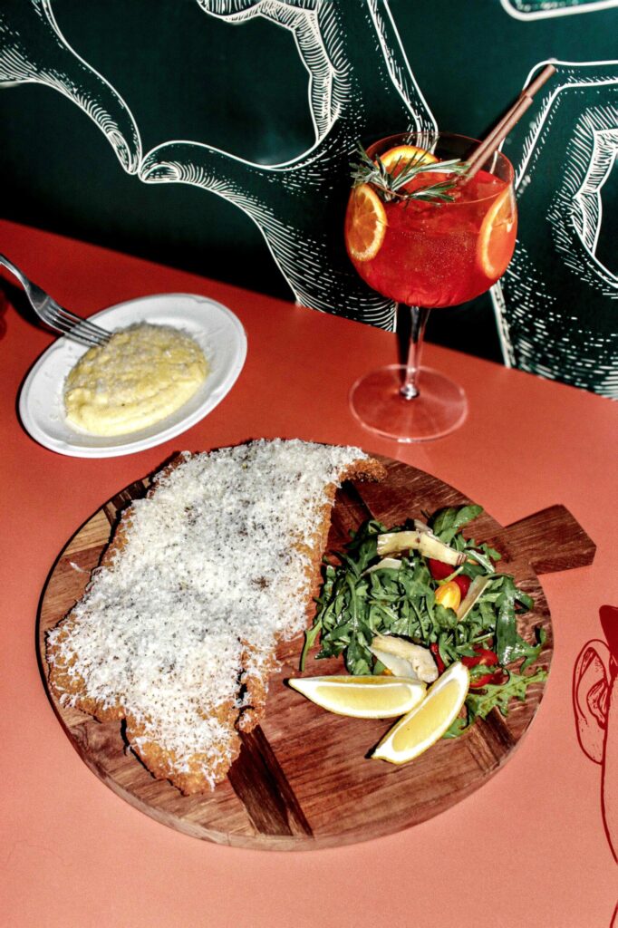 escalope panée façon milanaise, couverte de parmesan râpé, présentée sur une planche en bois avec salade (roquette, légumes) et quartiers de citron ; à côté, un cocktail rouge garni d’orange et de romarin, devant un mur illustré