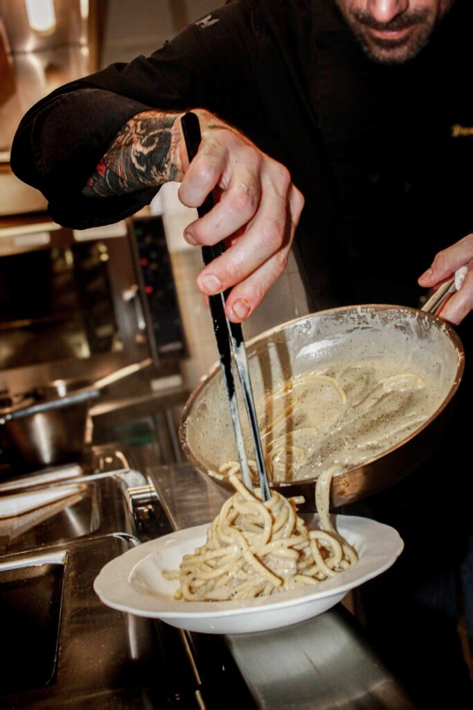 Photo en cuisine professionnelle : un chef en veste noire, bras tatoué, dresse des spaghettis nappés de sauce crémeuse (poivre visible) à l’aide d’une pince, depuis une poêle inclinée vers l’assiette.
