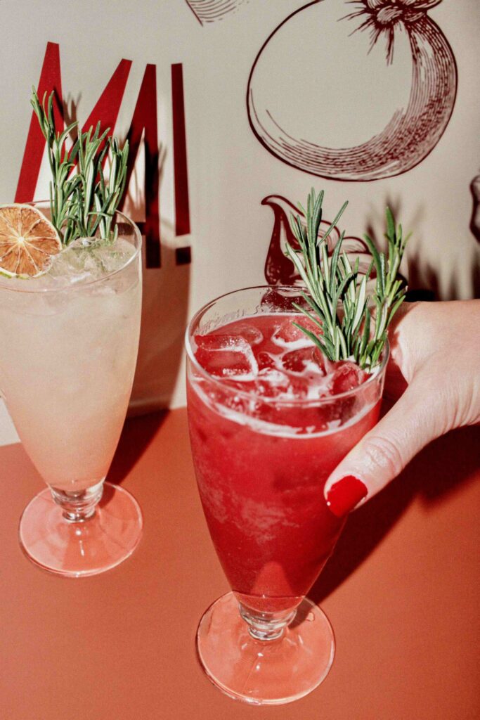 Photo en gros plan : deux cocktails sur une table, décorés de brins de romarin ; un cocktail rouge avec glaçons est tenu par une main aux ongles rouges, à côté d’un cocktail clair avec agrume, devant un décor mural illustré.