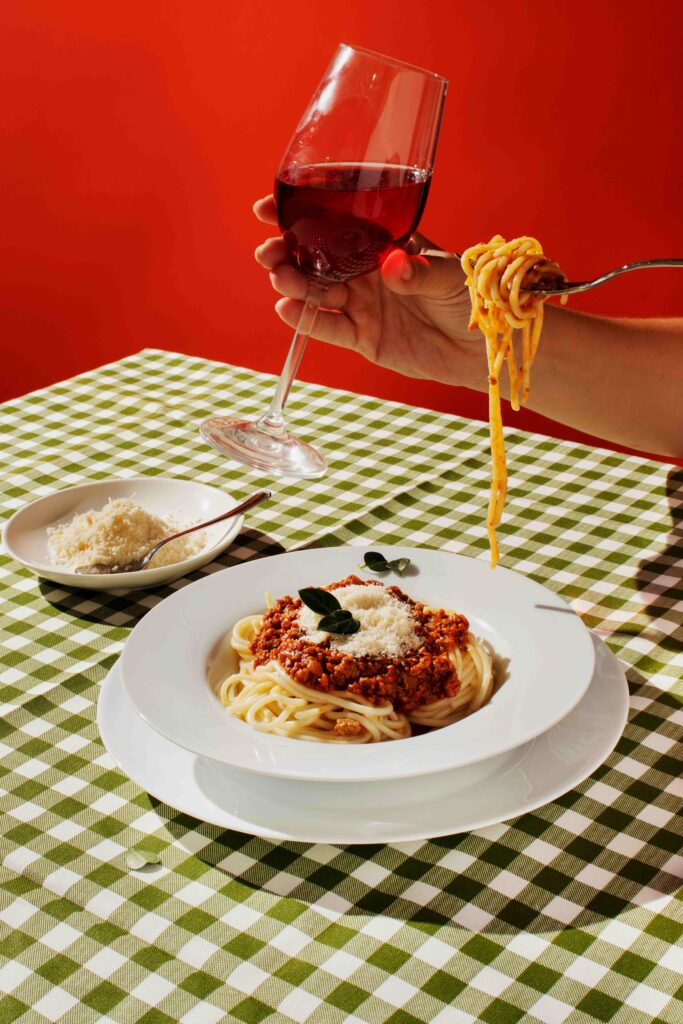 Scène de table : spaghetti bolognaise garnis de parmesan et basilic, bol de parmesan à côté, nappe vichy verte, fond rouge, une main lève un verre de vin rouge et une fourchette enroule des pâtes