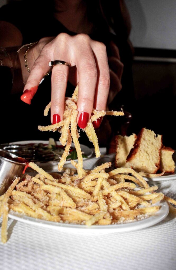 Photo en gros plan d’une table de restaurant : une main avec ongles rouges et bague attrape des pâtes frites/croustillantes saupoudrées de fromage, près d’un ramequin de sauce et de mor-ceaux de pain.