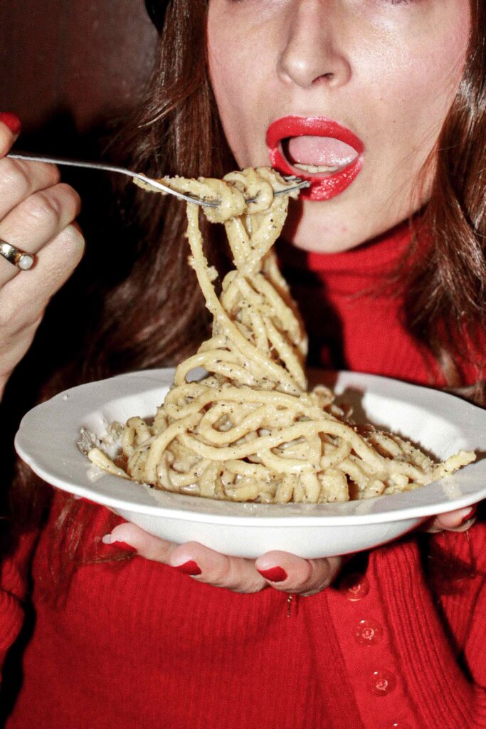 Photo en gros plan d’une personne habillée en rouge, avec lèvres et manucure rouges, tenant une assiette de spaghetti poivrés et enroulant les pâtes sur une fourchette.