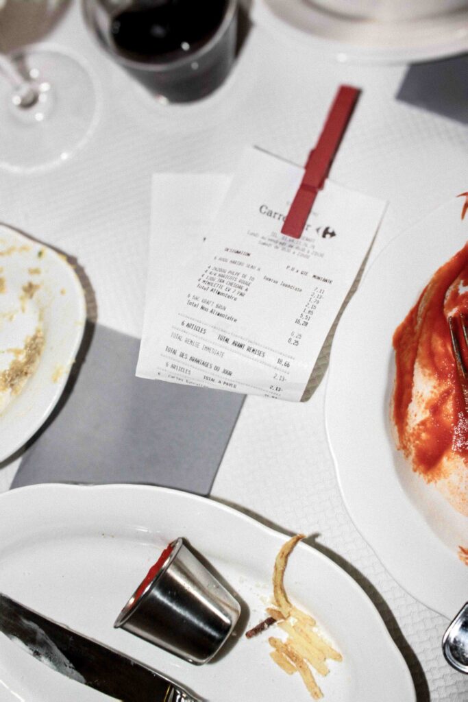 Photo en plongée d’une fin de repas : une addition maintenue par une pince rouge, un verre de vin rouge, des assiettes avec restes de pâtes et de sauce, et un petit ramequin renversé.