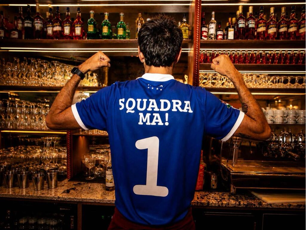 Vue de dos d'un membre de la Squadra MA! portant un maillot bleu numéro 1 devant le bar du Bouillon Chic Italien, dans une ambiance chaleureuse et conviviale.