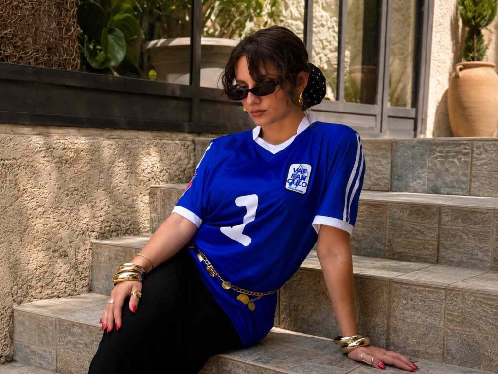 Mimaa artiste, jeune femme portant le maillot bleu VAF FAN CULO assise sur les marches d'une terrasse en extérieur, dans une ambiance lifestyle élégante et ensoleillée.