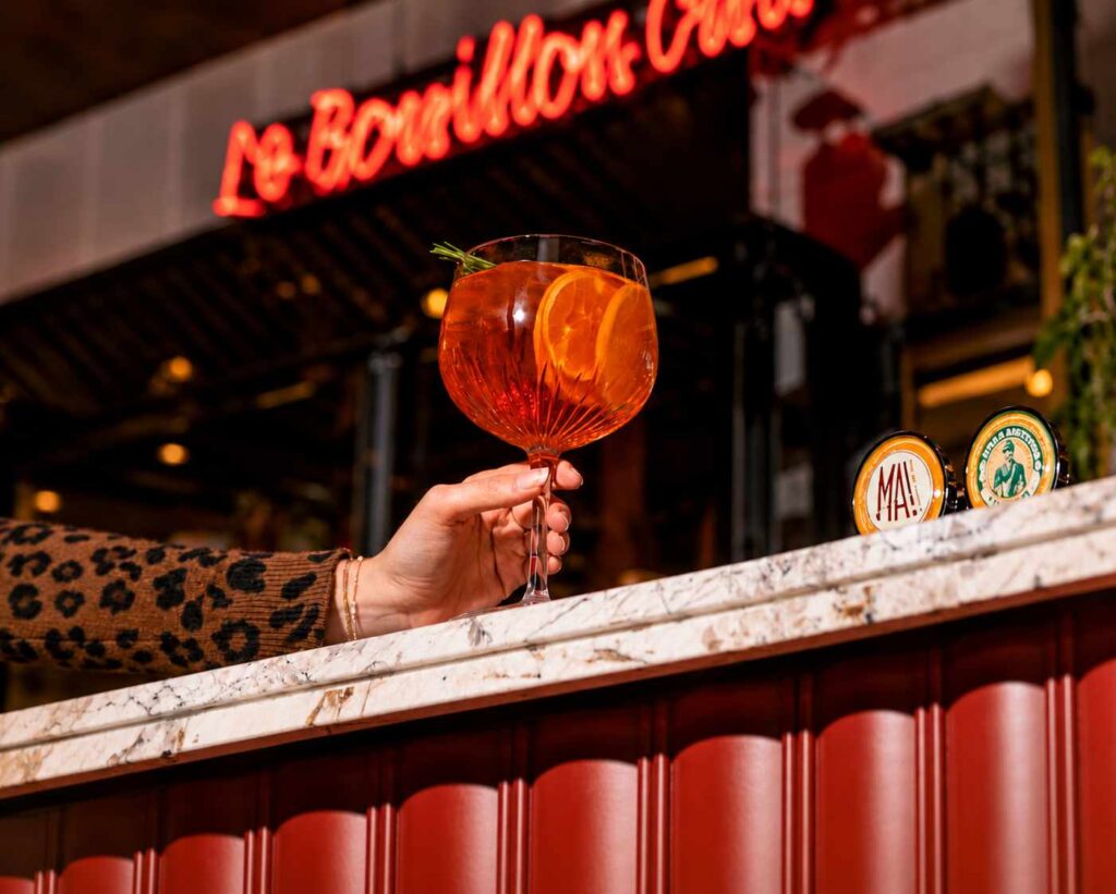 Cocktail orange garni d'une tranche d'orange et de romarin, servi au comptoir du Bouillon Caffè devant une enseigne lumineuse.