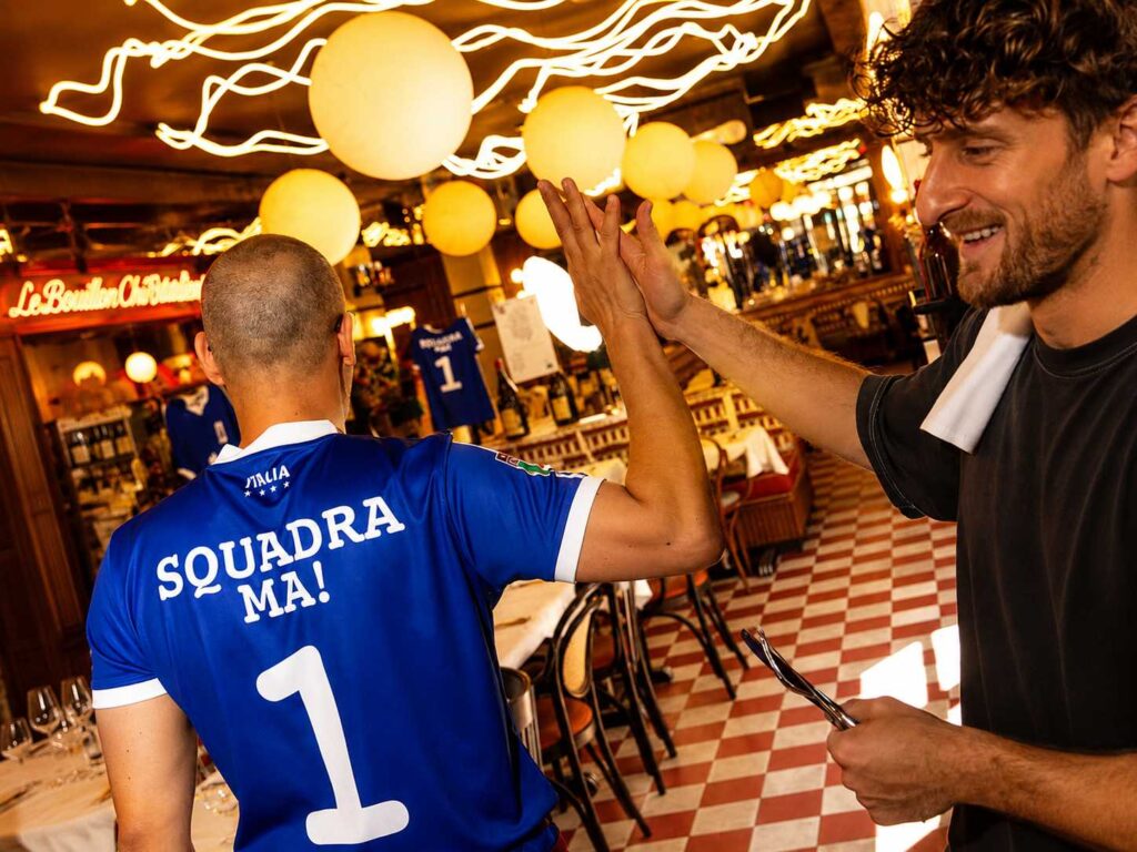 Deux membres de la Squadra MA! se tapent dans la main au cœur du restaurant, dans une ambiance chaleureuse et conviviale, devant la salle du Bouillon Chic Italien.