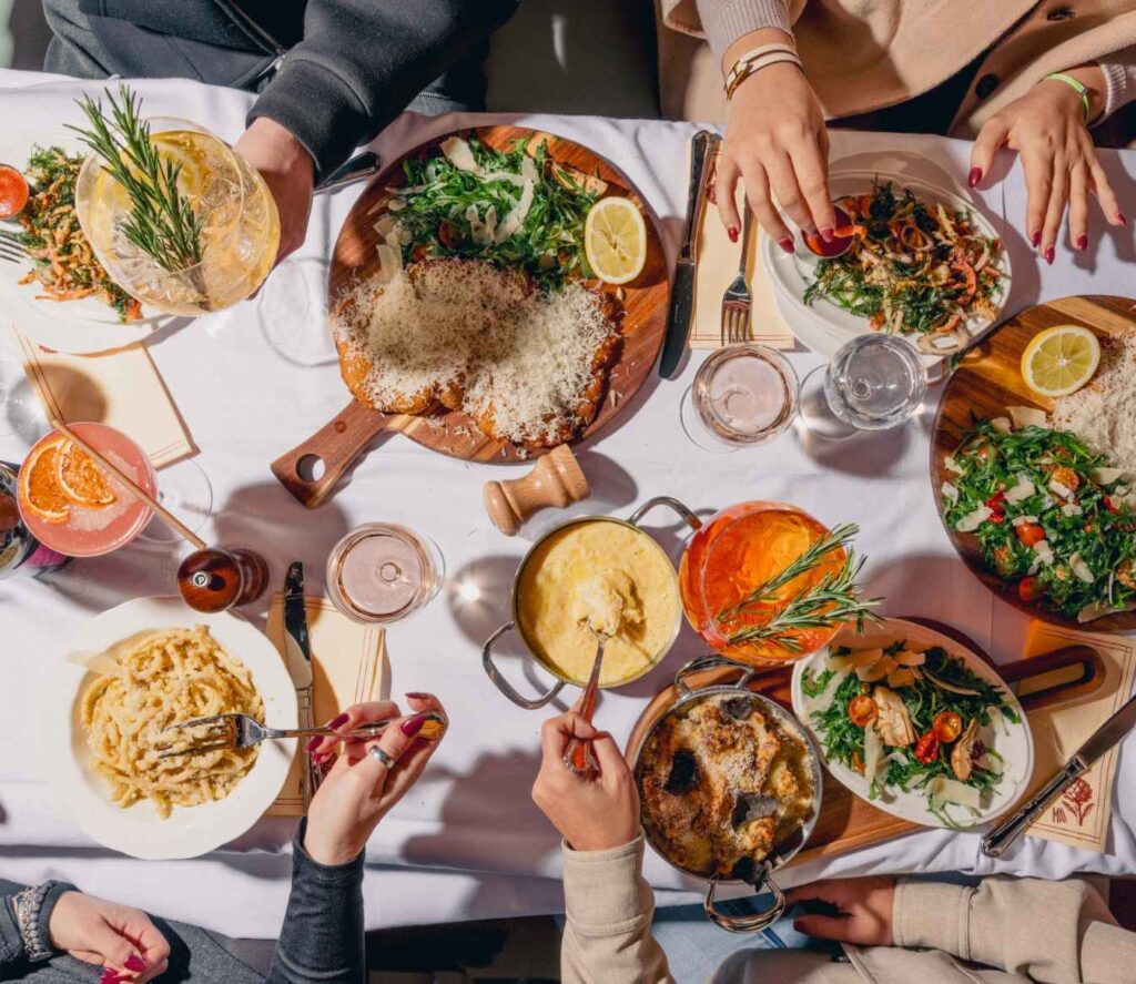 Vue de dessus d'une grande table conviviale garnie de spécialités italiennes à partager : escalopes milanaises, pâtes, salades, cocktails et antipasti au restaurant MA Bouillon.