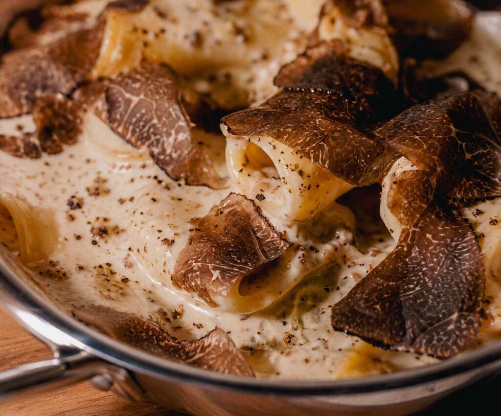 Gros plan sur des paccheri nappés d'une crème de parmesan et généreusement garnis de fines lamelles de truffe noire, servis dans une poêle en métal.