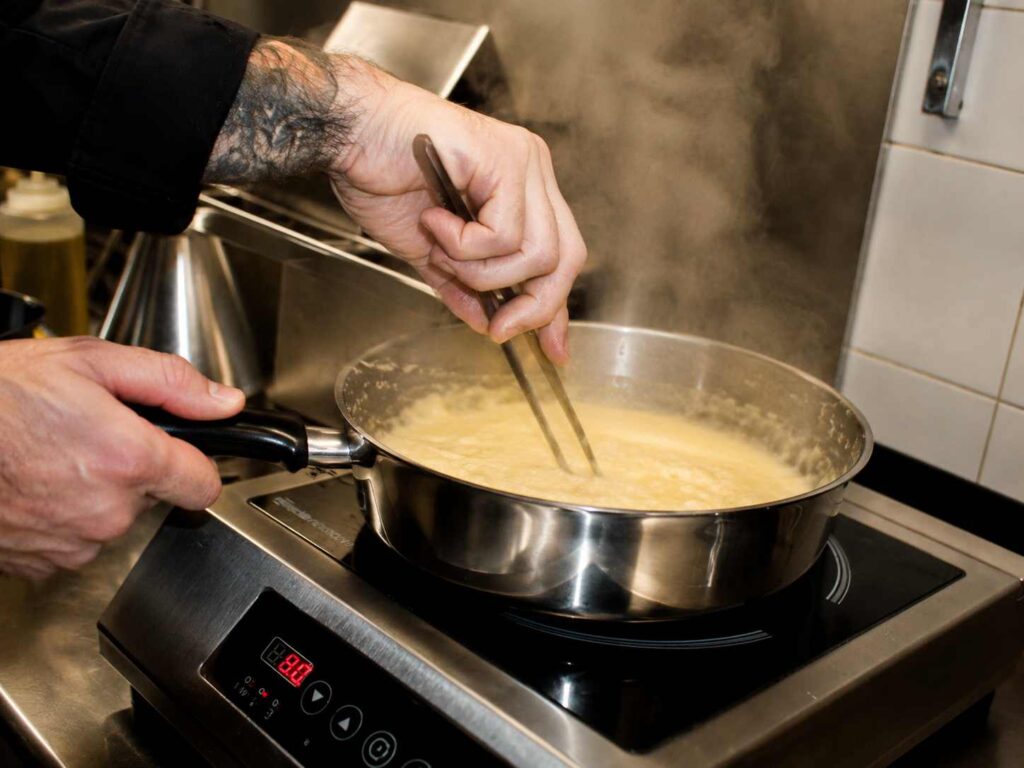 Chef préparant une sauce crémeuse pour des pâtes dans une poêle fumante sur une plaque à induction, en cuisine de restaurant italien