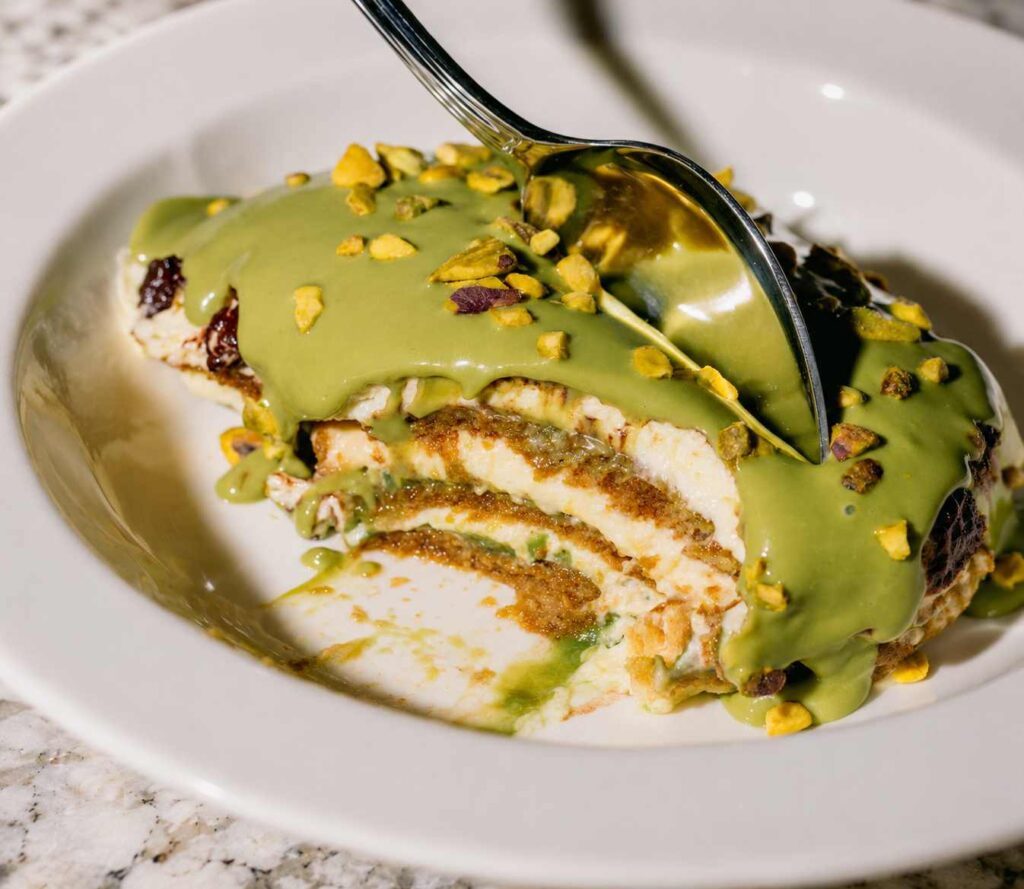 Part de tiramisu à la pistache nappée d'une généreuse crème de pistache et parsemée d'éclats de pistaches, servie dans une assiette blanche.
