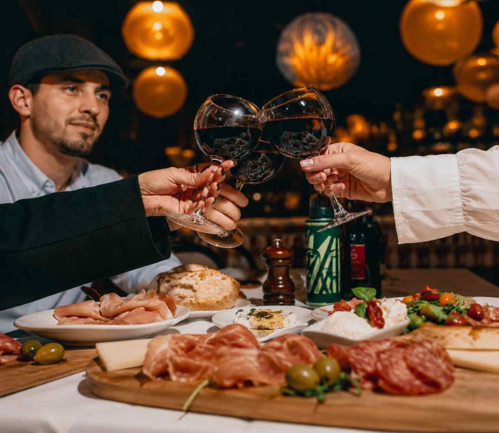 Convives trinquant avec des verres de vin rouge autour d'une table garnie d'antipasti, de charcuteries et de spécialités italiennes dans une ambiance chaleureuse