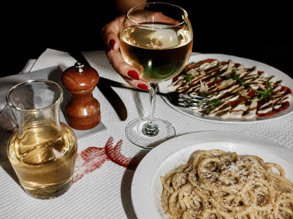 Une main tenant un verre de vin blanc au-dessus d'une table de restaurant italien avec une assiette de cacio e pepe, une salade caprese et une carafe de vin blanc