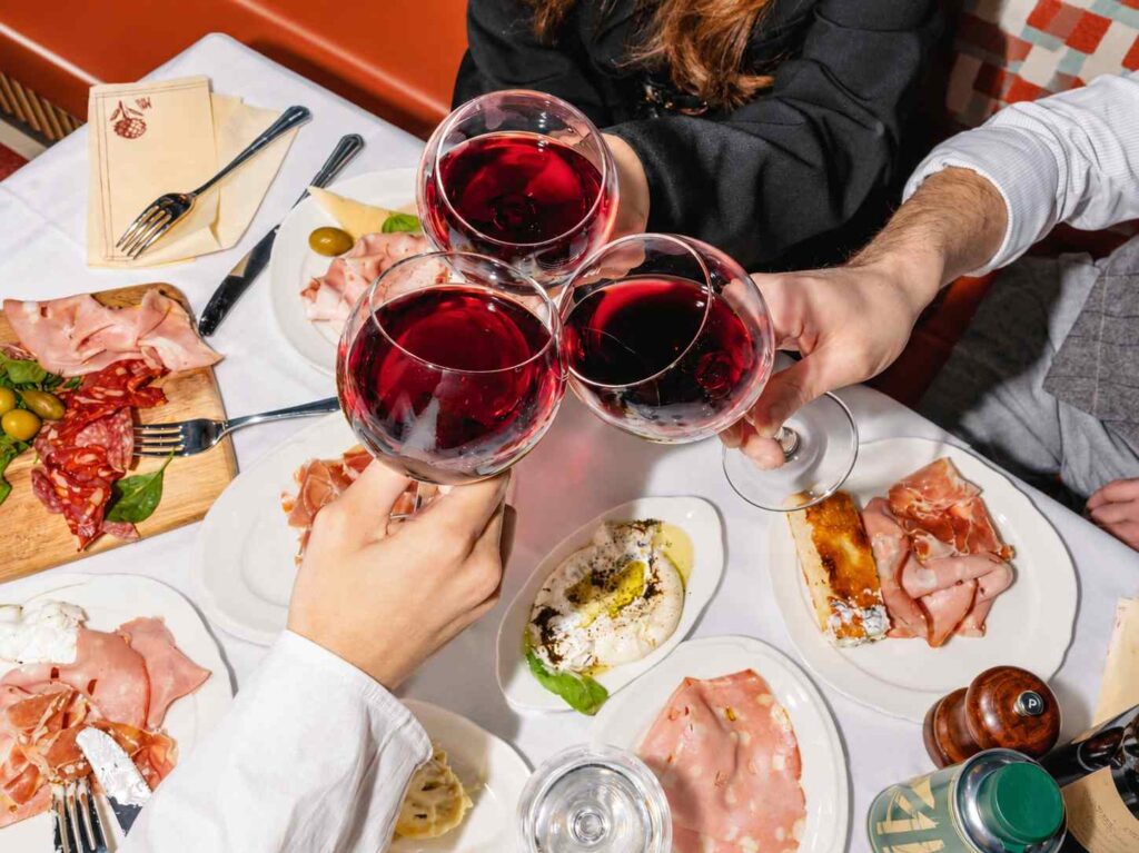 Trois personnes trinquent avec des verres de vin rouge autour d’une table garnie de charcuterie et de plats italiens.