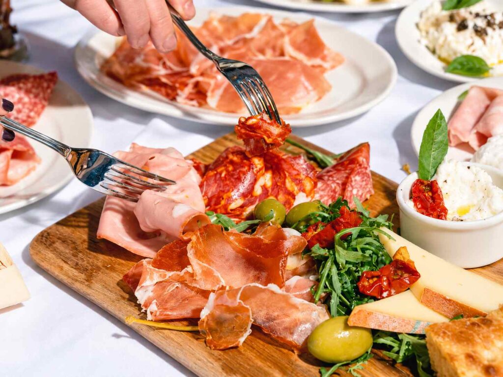 Des convives se servent avec leurs fourchettes sur une planche de charcuterie, fromages et olives, accompagnée de burrata.