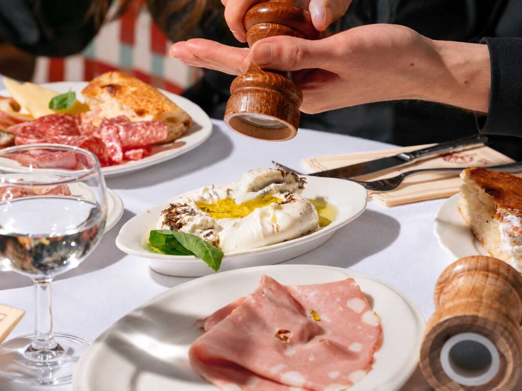 Une personne tient un moulin à poivre au-dessus d’une burrata à l’huile d’olive, entourée d’assiettes de charcuterie et de pain.