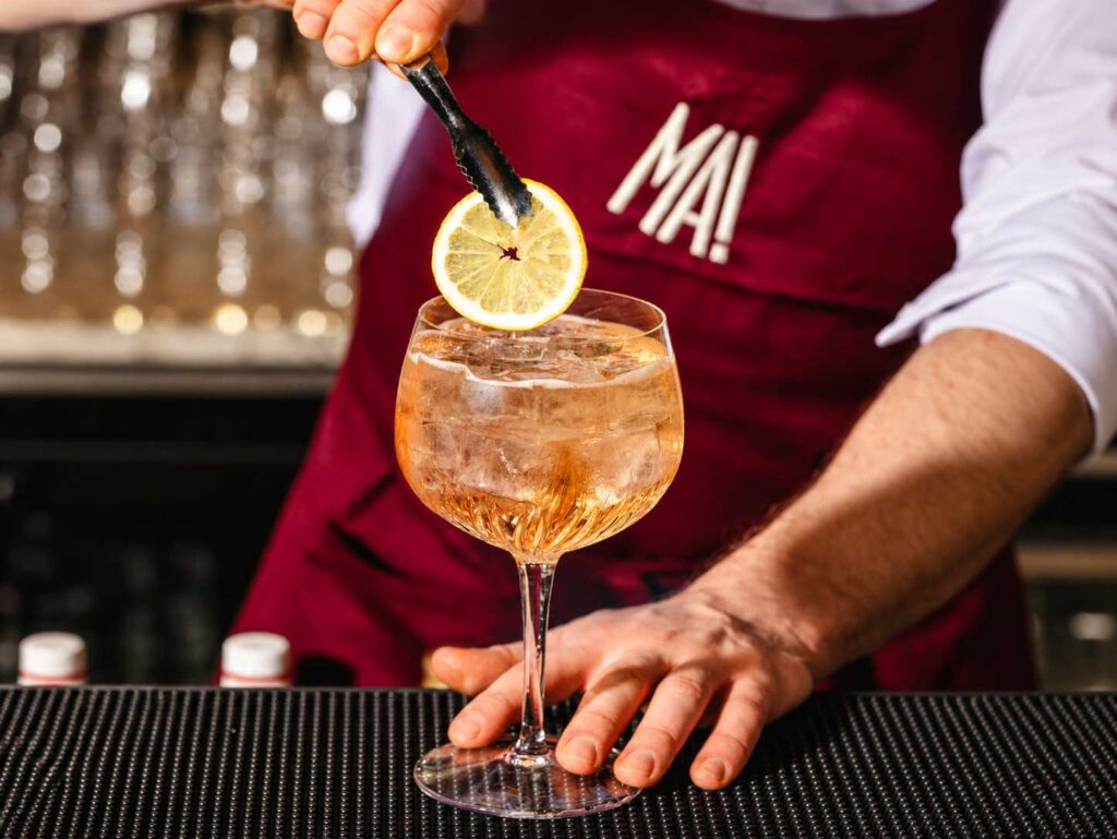 Un barman dépose une rondelle de citron sur un cocktail ambré rempli de glaçons, au bar de MA! Le Bouillon Chic Italien.