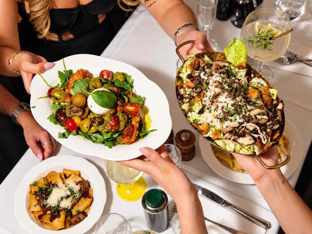 Des convives se passent une salade à la burrata et un grand plat de salade, avec une assiette de pâtes sur la table.