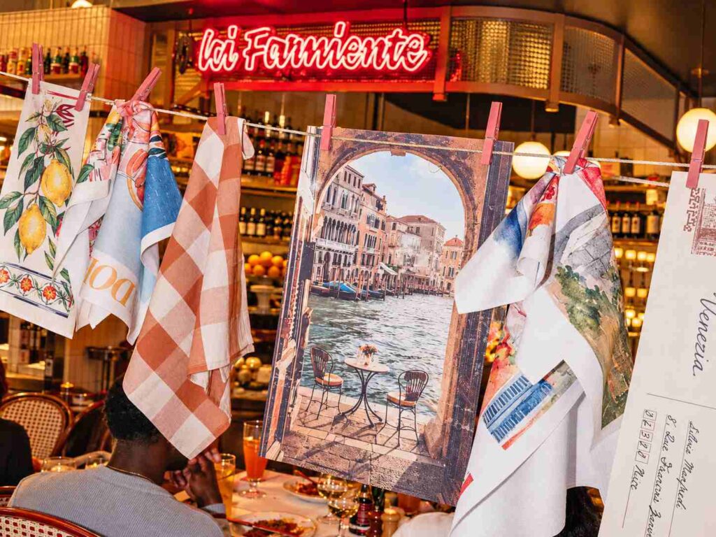 Des tissus et illustrations d’inspiration italienne sont suspendus par des pinces roses dans la salle du restaurant, sous un néon rouge.